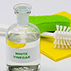 Glass bottle of white vinegar with a scrubbing brush, green sponge, and yellow cloth in the background