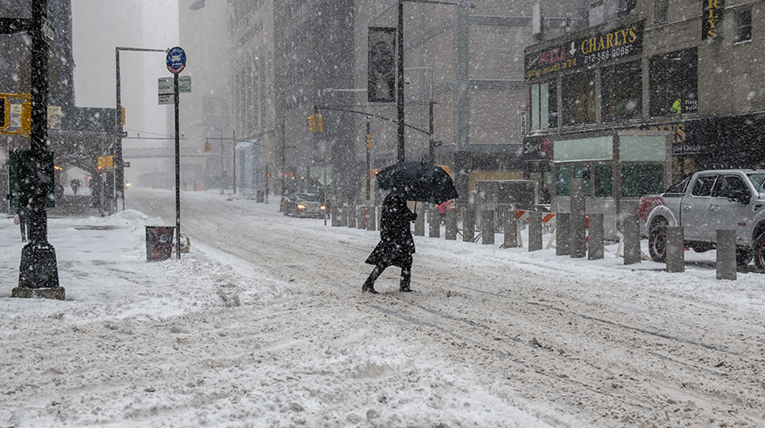 City street covered in snow and a man in a black jacket walking by with an umbrella