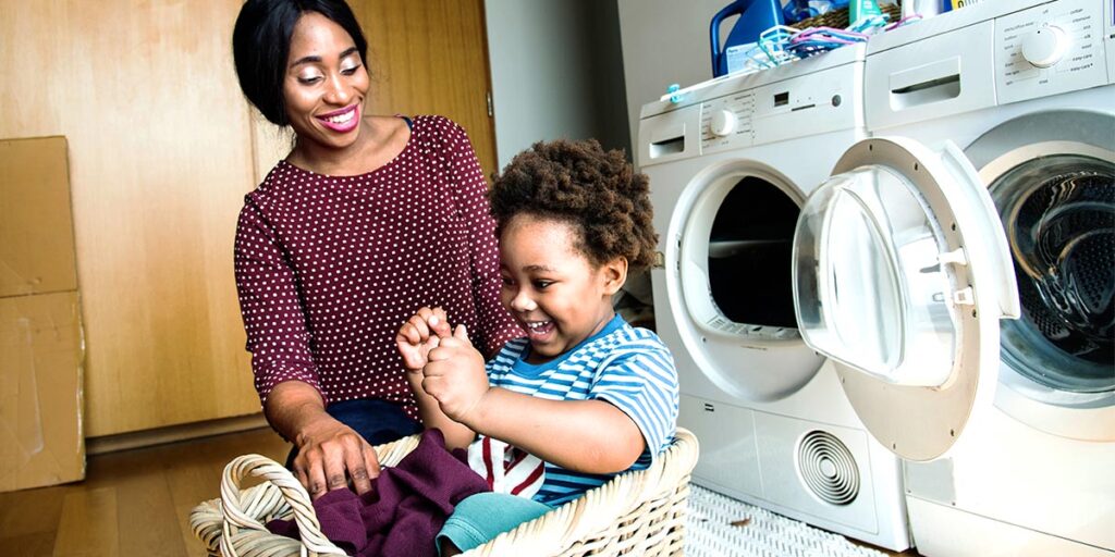 In a laundry room, a woman smiling at her toddler son, who sits giggling in a laundry basket.